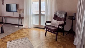 A living room with a leather armchair, a television, a wooden table, and a large window with white curtains.