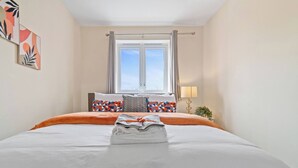 A neatly made bed with a folded towel, colorful pillows, and a window with curtains. - 3 Bedroom Cosy-home in Hornchurch With hot tub (Hornchurch)