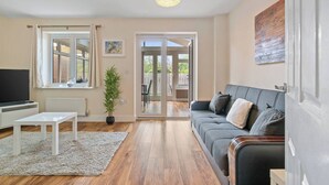 A modern living room with a sofa, a TV, a white coffee table, and a large window. - 3 Bedroom Cosy-home in Hornchurch With hot tub (Hornchurch)