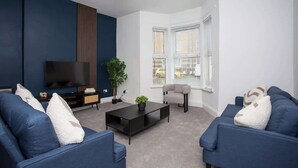 Uma sala de estar moderna com televisão de tela plana, sofás azul escuro, uma mesa de centro e um vaso com planta.