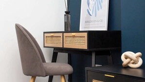 Quarto moderno com parede azul escura, uma cadeira cinza, escrivaninha preta com detalhes de rattan e uma obra de arte emoldurada.