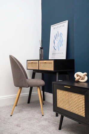 Ein modernes Zimmer mit einer dunkelblauen Wand, einem grauen Sessel, einem schwarzen Schreibtisch mit Rattan-Details und einem gerahmten Kunstwerk.