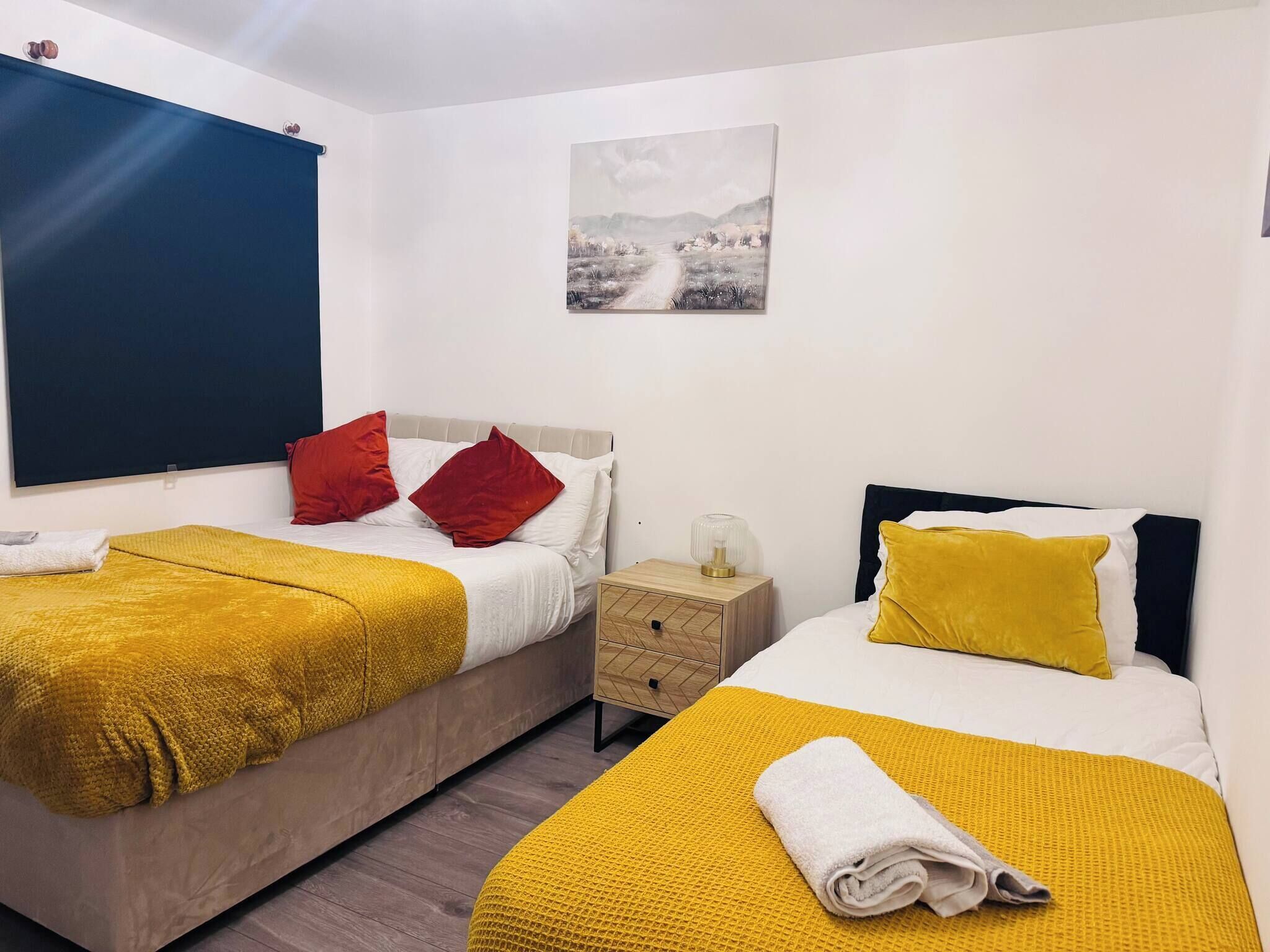 Habitación de hotel con dos camas, cada una con una manta y almohadas amarillas, una mesita de noche con lámpara y un cuadro en la pared.