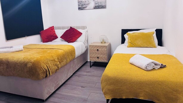 Een hotelkamer met twee bedden, elk met een gele deken, witte beddengoed en rode en gele kussens. Er is een klein nachtkastje met een lamp, een kunstwerk aan de muur en een raam met een blauwe rolgordijn.