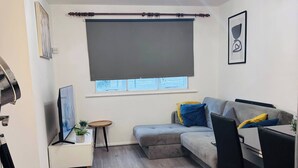 Een moderne woonkamer met een grijze bank, een eettafel met stoelen, een klein bijzettafeltje met een plant, een groot raam met een donker gordijn en een plafondlamp.