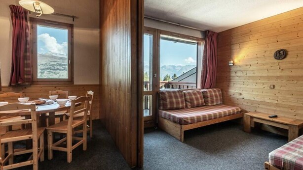 A cozy room with a wooden interior, a dining table with chairs, a sofa, and a view of mountains through large windows. - ZIG ZAG 39 (La Plagne-Tarentaise)