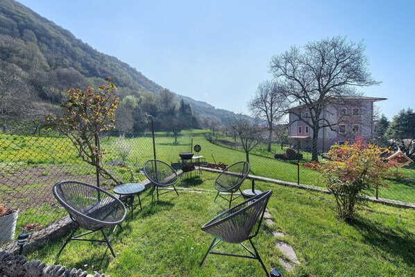 A fenced outdoor area with chairs and a table, a building in the background, and a hilly landscape.
