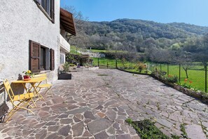 A stone patio with a table and chairs, a house with shutters, and a view of a green field and hills.