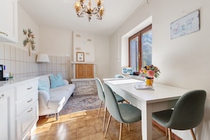 A modern kitchen with a dining area, white cabinets, a chandelier, and a view through a window.