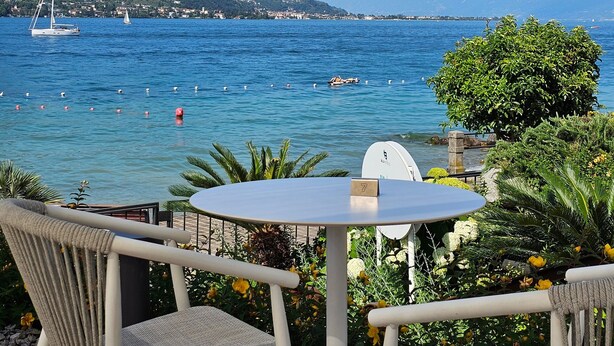 Une table et deux chaises sont placées face à un plan d’eau, avec des bateaux et des montagnes en arrière-plan.