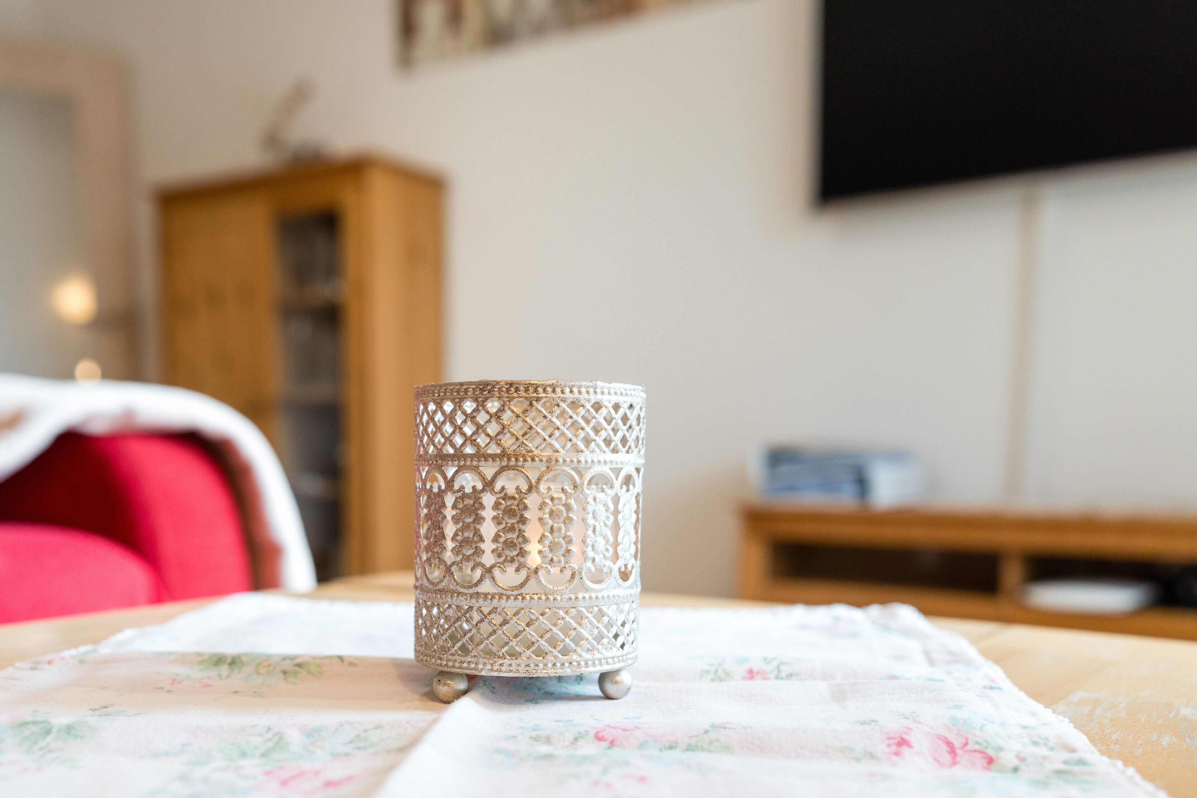 A decorative candle holder on a floral tablecloth in a room with a wooden cabinet and a television in the background.