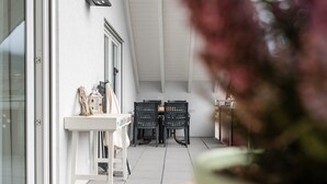 Un patio con una mesa y sillas blancas, un pequeño estante con adornos, y vista al interior a través de una puerta de vidrio.