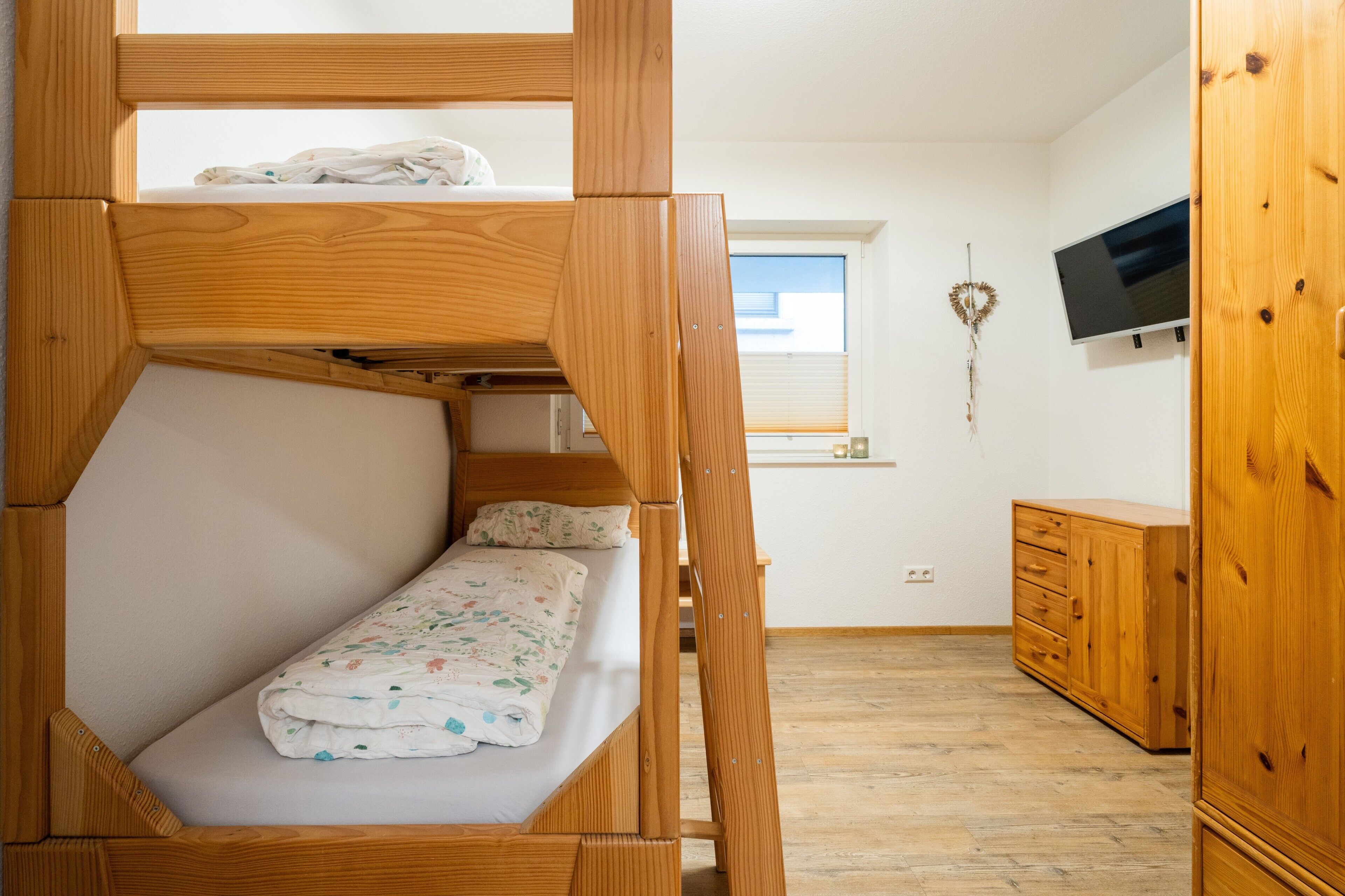A bunk bed with a wooden frame, a small bedside table, a wardrobe, and a television on the wall.