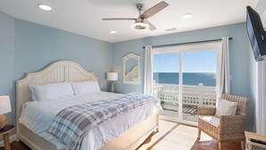 Une chambre à coucher comprenant un lit, un ventilateur de plafond, une table de chevet, une lampe, un fauteuil en osier, un téléviseur et un balcon avec vue sur l’océan.