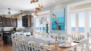 Une cuisine moderne avec des meubles de couleur sombre, un coin repas avec des chaises blanches et une table en bois, et un grand tableau représentant un oiseau perché sur un poteau.