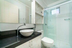 A modern bathroom with a white sink, black countertop, and a glass-enclosed shower. - Apartamento 2 quartos vista mar Bombas (Bombinhas)