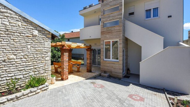 A modern two-story house with a stone wall, a covered patio with a bench, and a paved courtyard. - Sea View Villa Near Beach in Posedarje (Posedarje)