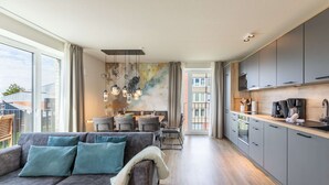 A modern living room with a grey sofa, dining area with a table and chairs, and a kitchen with grey cabinets and a wooden countertop. - Luxusappartement auf Nordstrand (Nordstrand)