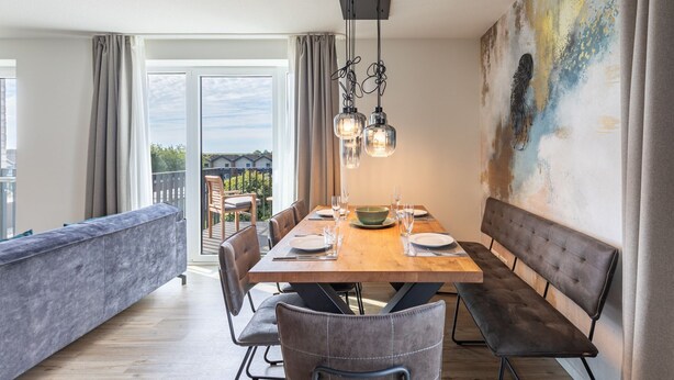 A dining area with a wooden table set for four, surrounded by grey upholstered chairs. A bench is positioned along the wall, and a large abstract painting is visible. - Luxusappartement auf Nordstrand (Nordstrand)