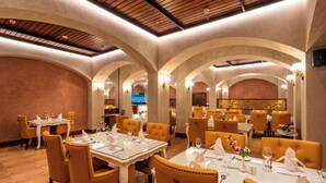 A restaurant with high ceilings, arched alcoves, and a warm color palette. Tables are set with white tablecloths and gold chairs. - Jajati Courtyard (Korai)