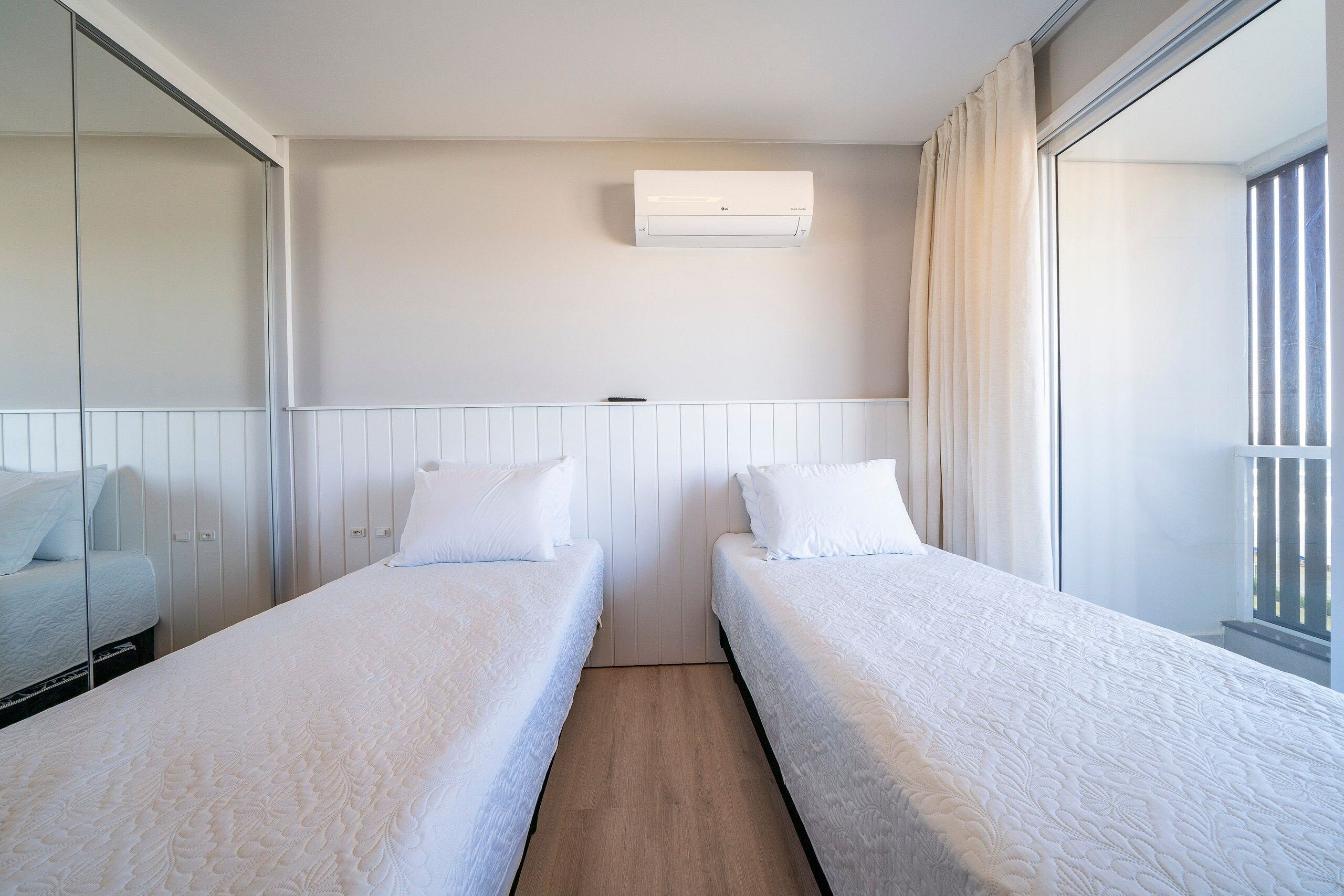 Hotellihuone, jossa on kaksi erillistä sänkyä, seinään asennettu ilmastointilaite, ikkuna verhoilla ja peilillä varustettu vaatekaappi.