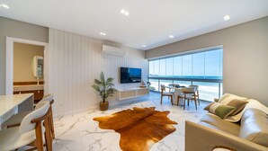 A modern living room with a large window, a flat-screen TV, a leather sofa, and a dining area with chairs. - Duplex Alto Padrão Frente Mar c Piscina (Bombinhas)