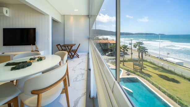 A modern room with a large window overlooking a beach and ocean. It includes a dining area with a table and chairs, a flat-screen TV, and a pool visible through the window. - Duplex Alto Padrão Frente Mar c Piscina (Bombinhas)