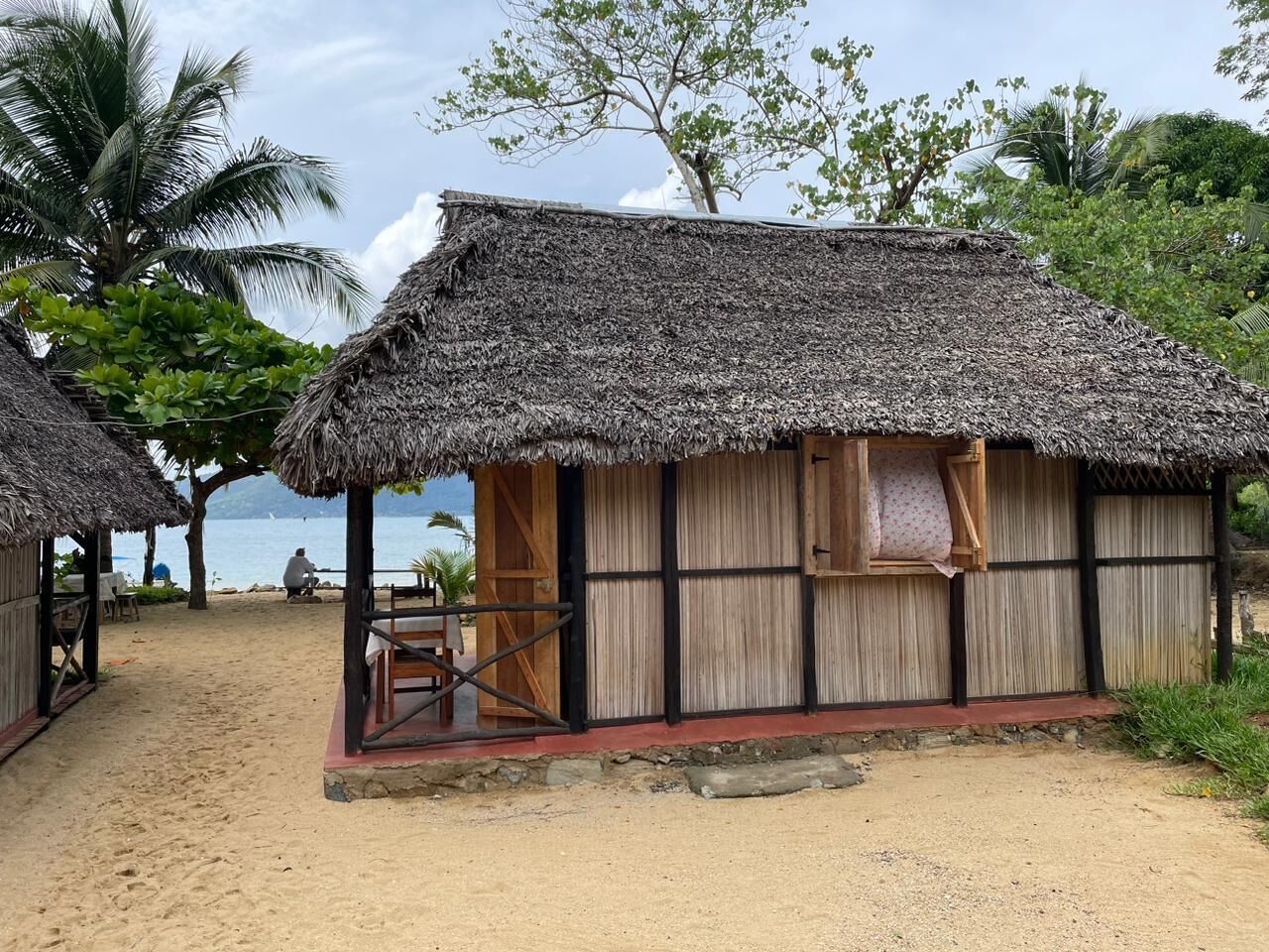 A thatched-roof hut with a wooden door and window, situated on a sandy beach with palm trees and a view of the sea in the background.