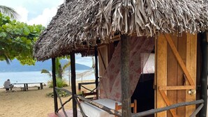 A wooden structure with a thatched roof, a bed, and a bench on a sandy beach with a view of the sea. - Djema Forest (Nosy Be)