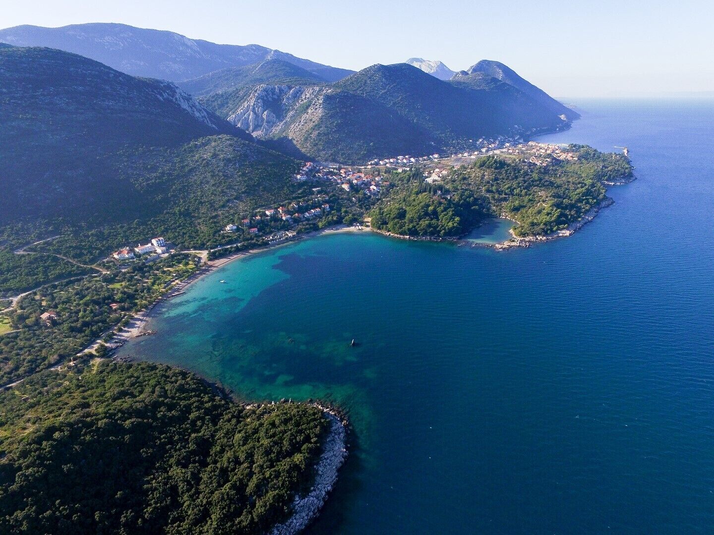 Une vue aérienne d’une ville côtière, avec une mer bleue et limpide, entourée de collines et de montagnes verdoyantes.