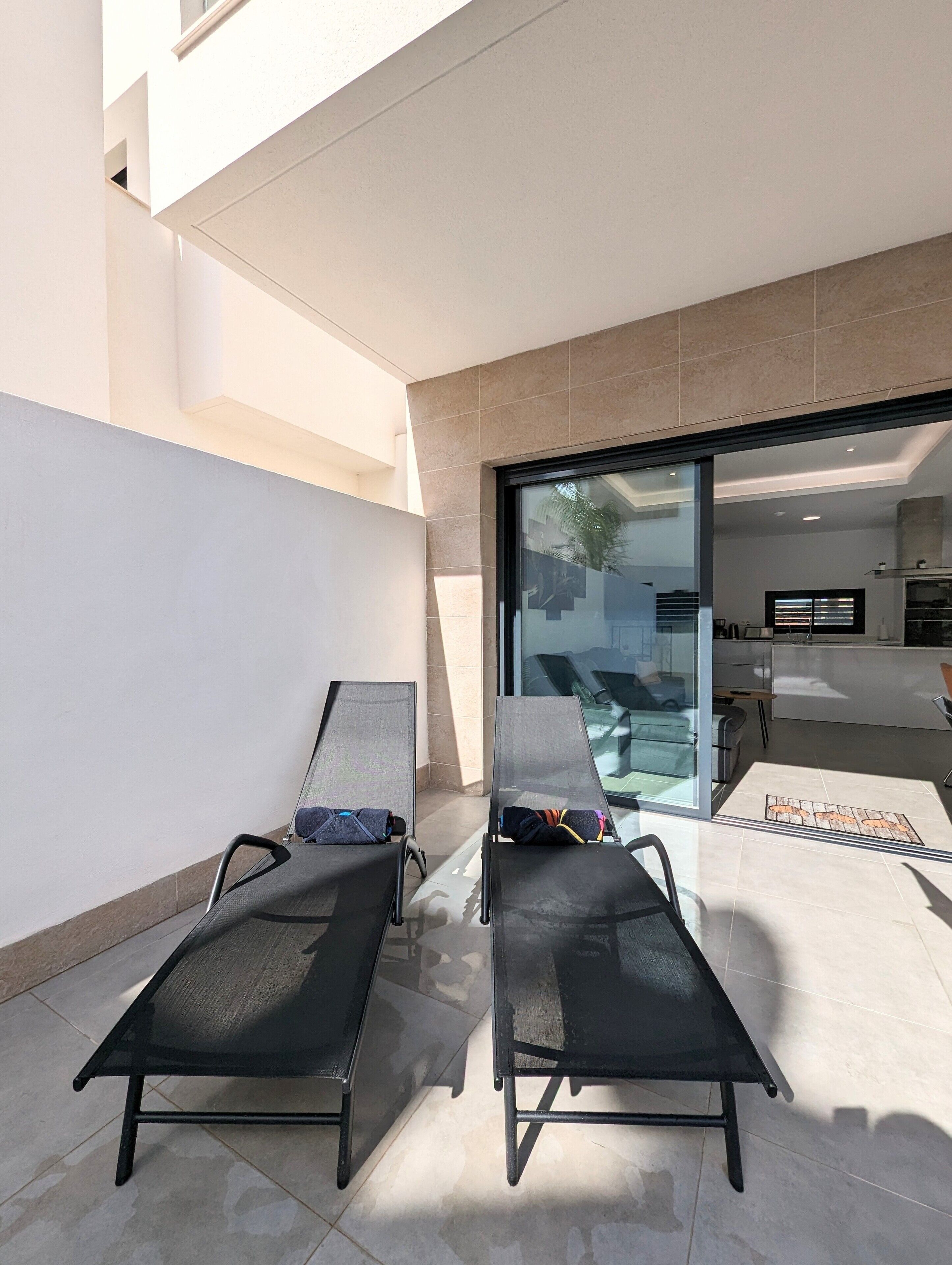 A modern outdoor seating area with two lounge chairs, a glass door leading to a pool, and a tiled floor.