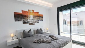 A modern bedroom with a large bed, a nightstand with a lamp, a wall-mounted artwork, and a view through a large window. - Villa 'R-58 Luxury New Build' with Private Pool, Terrace and Wi-Fi (Los Montesinos)