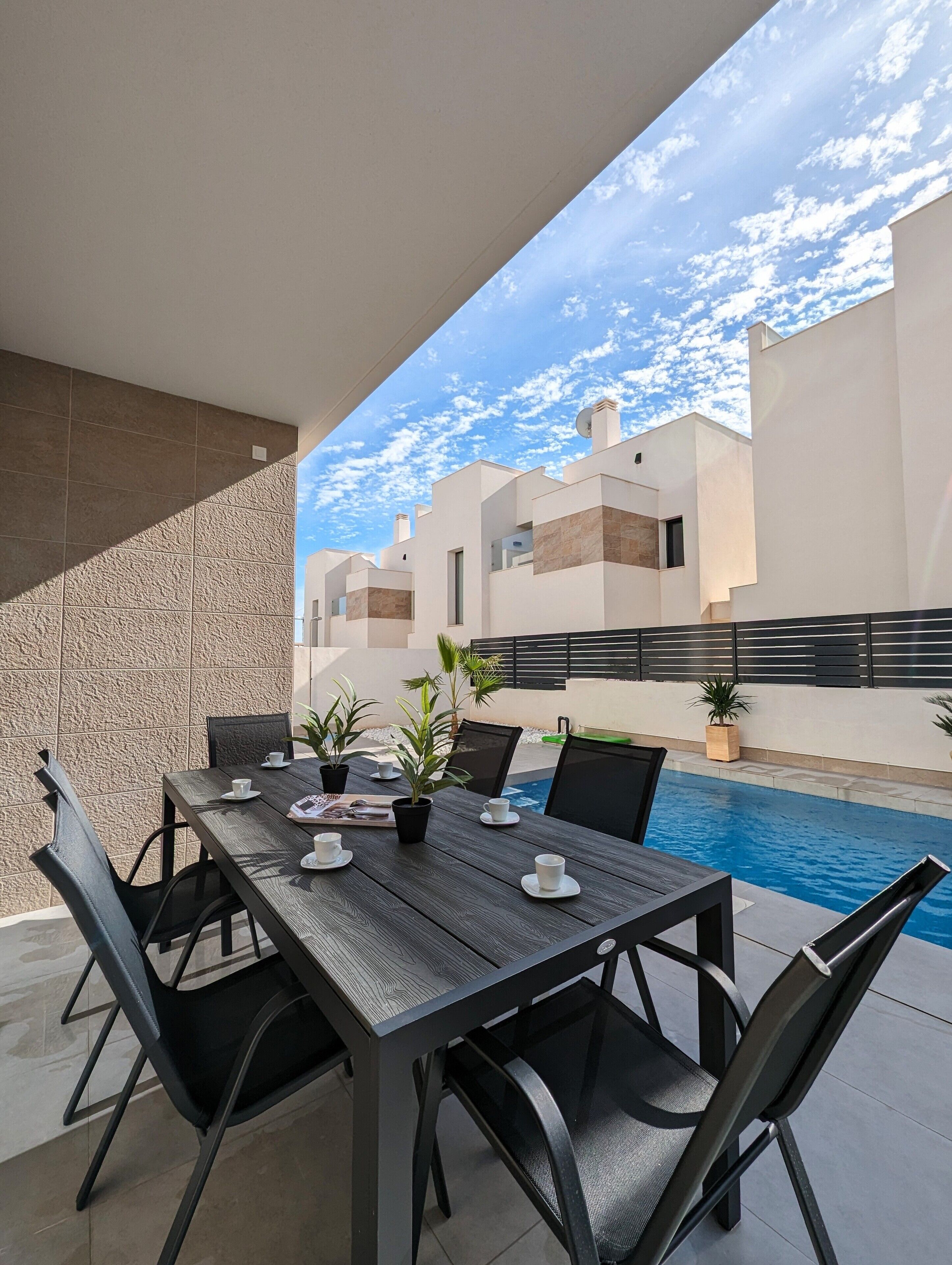 An outdoor patio with a dining table, chairs, potted plants, and a swimming pool in the background.