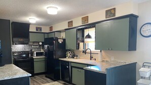 A kitchen with dark green cabinets, a black refrigerator, a microwave, and a sink. There is a clock on the wall and a window with blinds. - Rustic Ranch Retreat, Farm stay, Firepit, Game Room, Stars (Pauls Valley)