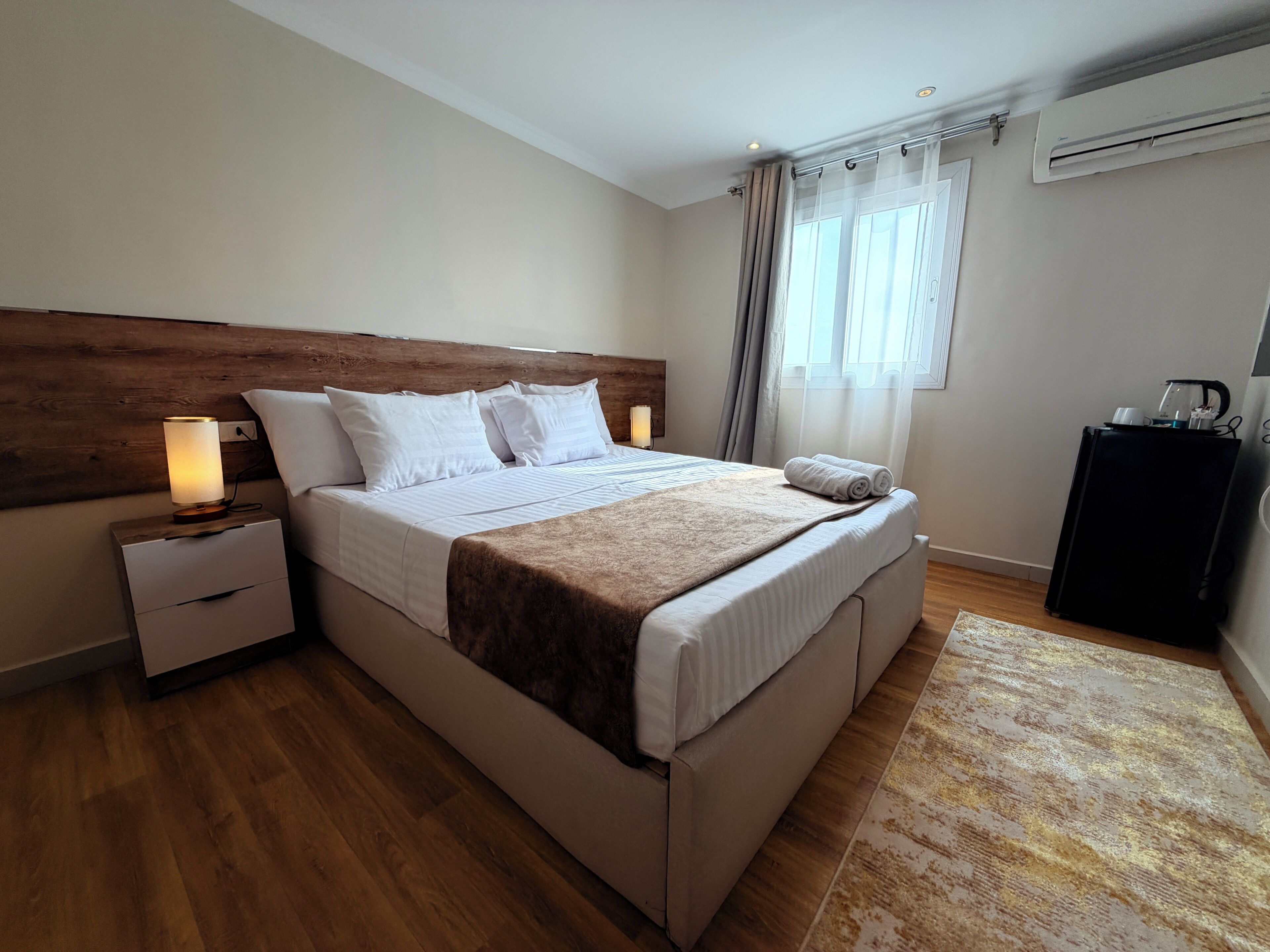 A bedroom with a wooden headboard, a bed with white linens, a bedside table with a lamp, a rolled towel, and a coffee machine on a desk.