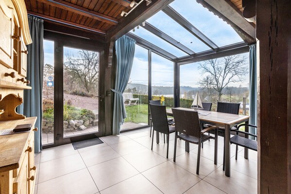 A room with a wooden cabinet, a dining table with chairs, and a large glass door leading to an outdoor area with a view of trees and hills.