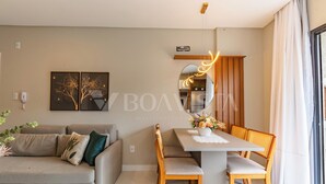 A modern living room with a sofa, dining table, and chairs. There is a decorative mirror above the table and a plant on the floor. - Sunny Flats 103 - Piscina Bombinhas (Bombinhas)