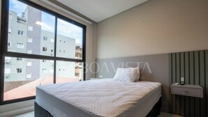 A modern bedroom with a large window, a bed with white bedding, a bedside table, and a view of a building outside. - Sunny Flats 103 - Piscina Bombinhas (Bombinhas)
