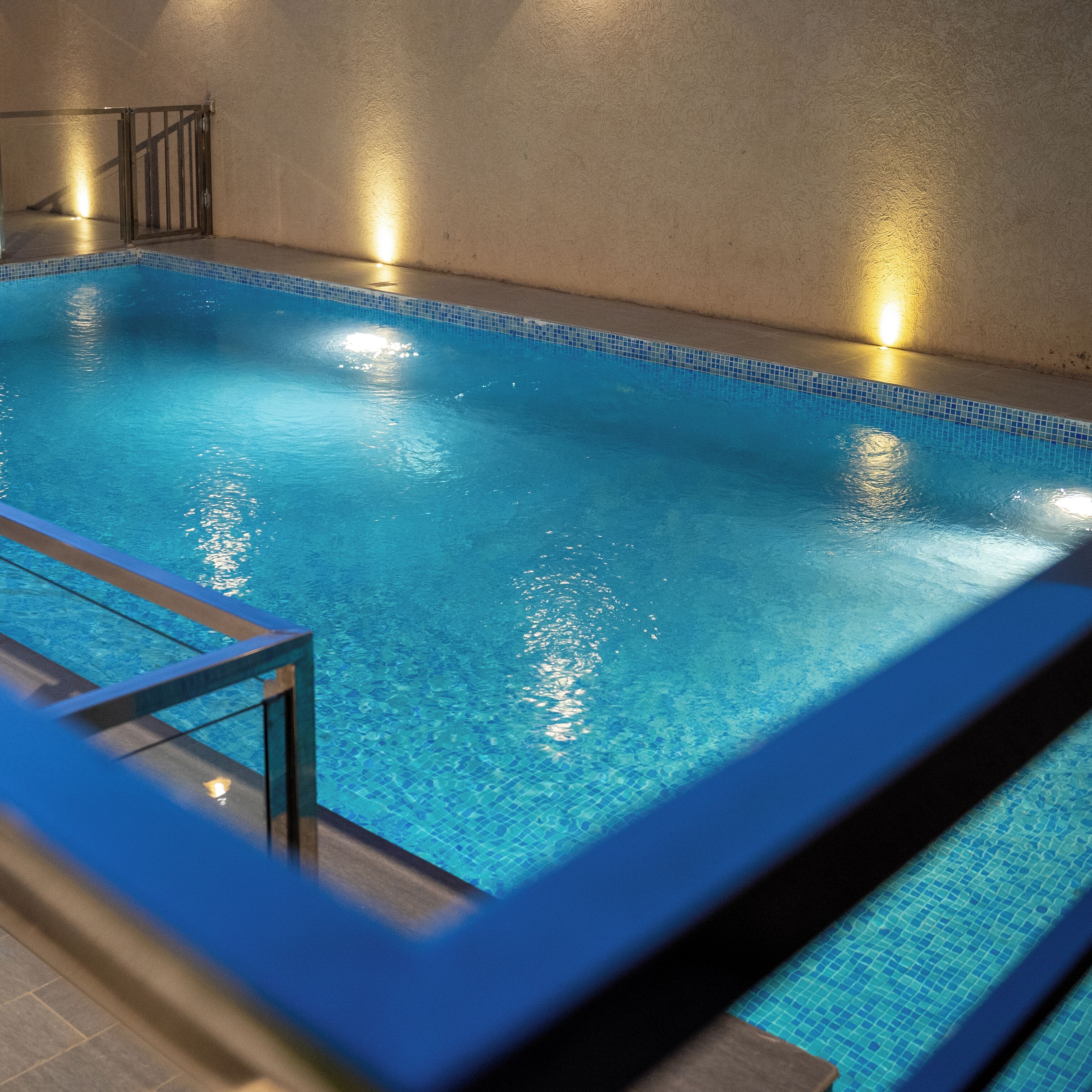 A rectangular indoor swimming pool with clear blue water, tiled edges, and a metal safety railing.