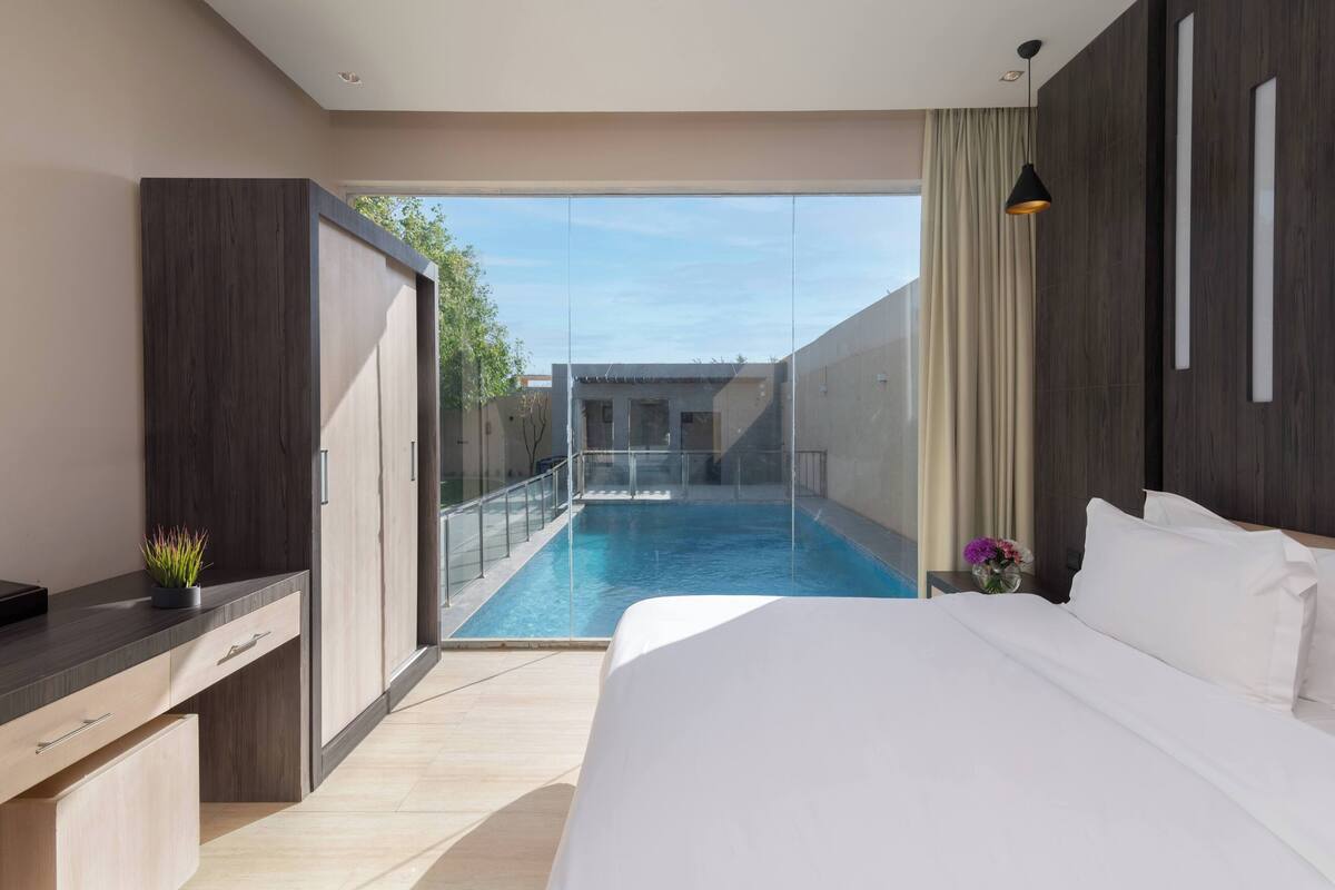 Une chambre d’hôtel moderne avec un grand lit, un bureau avec des tiroirs, une petite plante et une vue sur une piscine extérieure.