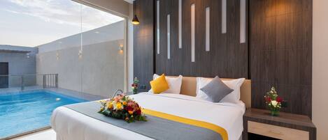 A modern hotel room with a large bed, a bedside table, a nightstand, and a view of an indoor swimming pool.