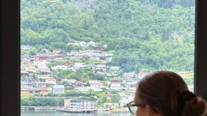 A person seated by a window overlooking a lake with buildings on the opposite shore.