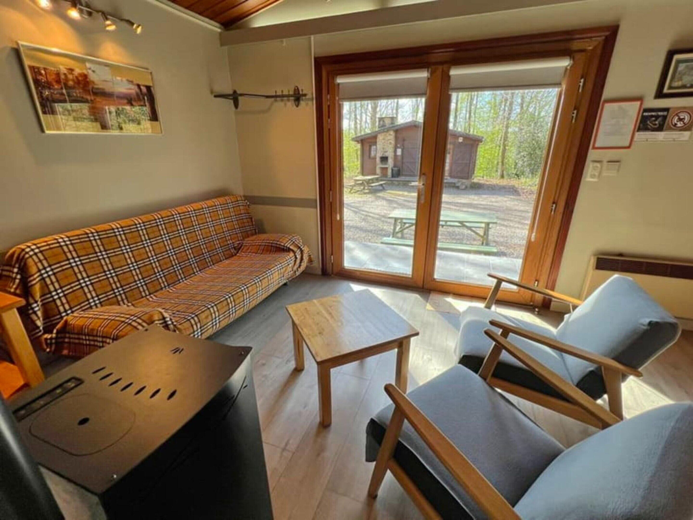 A cozy living area with a plaid sofa, a small table, and two chairs. A large window offers a view of an outdoor patio with a wooden structure.