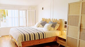 Un dormitorio con cama de madera, ropa de cama a rayas, una mesita de noche con lámpara y vistas al exterior a través de una ventana.