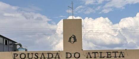 A sign for "POUSADA DO ATLETA" with a logo, a building in the background, and a hedge in the foreground.