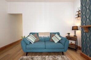 Ein smaragdgrünes Sofa mit gemusterten Kissen, ein kleiner Holztisch mit einer Lampe und eine Wand mit Muster.