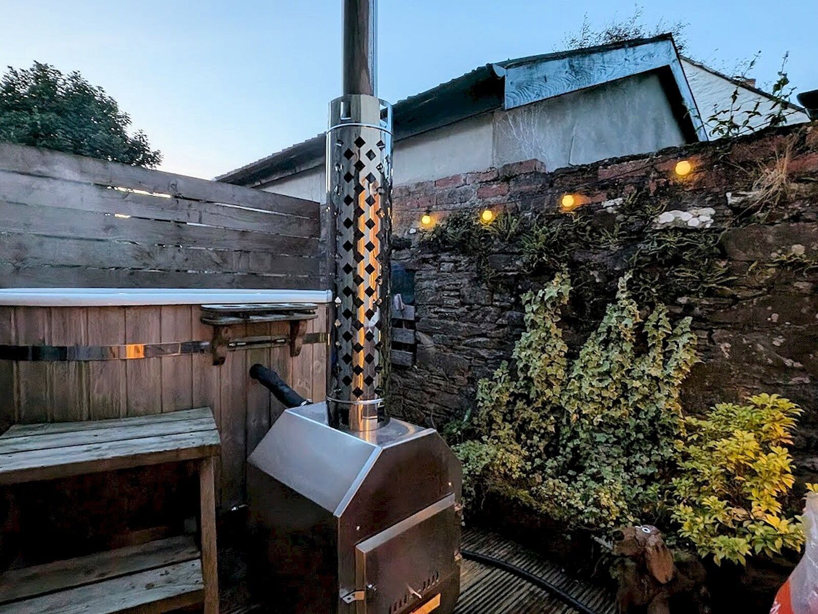 Una parrilla para barbacoa al aire libre con chimenea, un banco de madera y un muro de piedra con plantas.