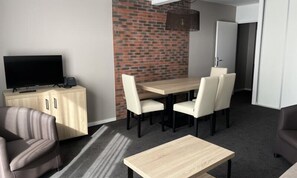 Ein modernes Hotelzimmer mit einem Flachbildfernseher, einem Holzkabinett, einem Esstisch mit vier Stühlen und einer Wand mit Backsteinverkleidung.