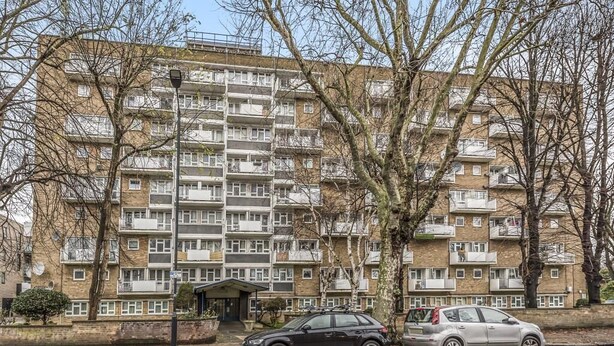Un edificio de apartamentos de varios pisos con balcones, un estacionamiento y una pequeña entrada con porche cubierto.