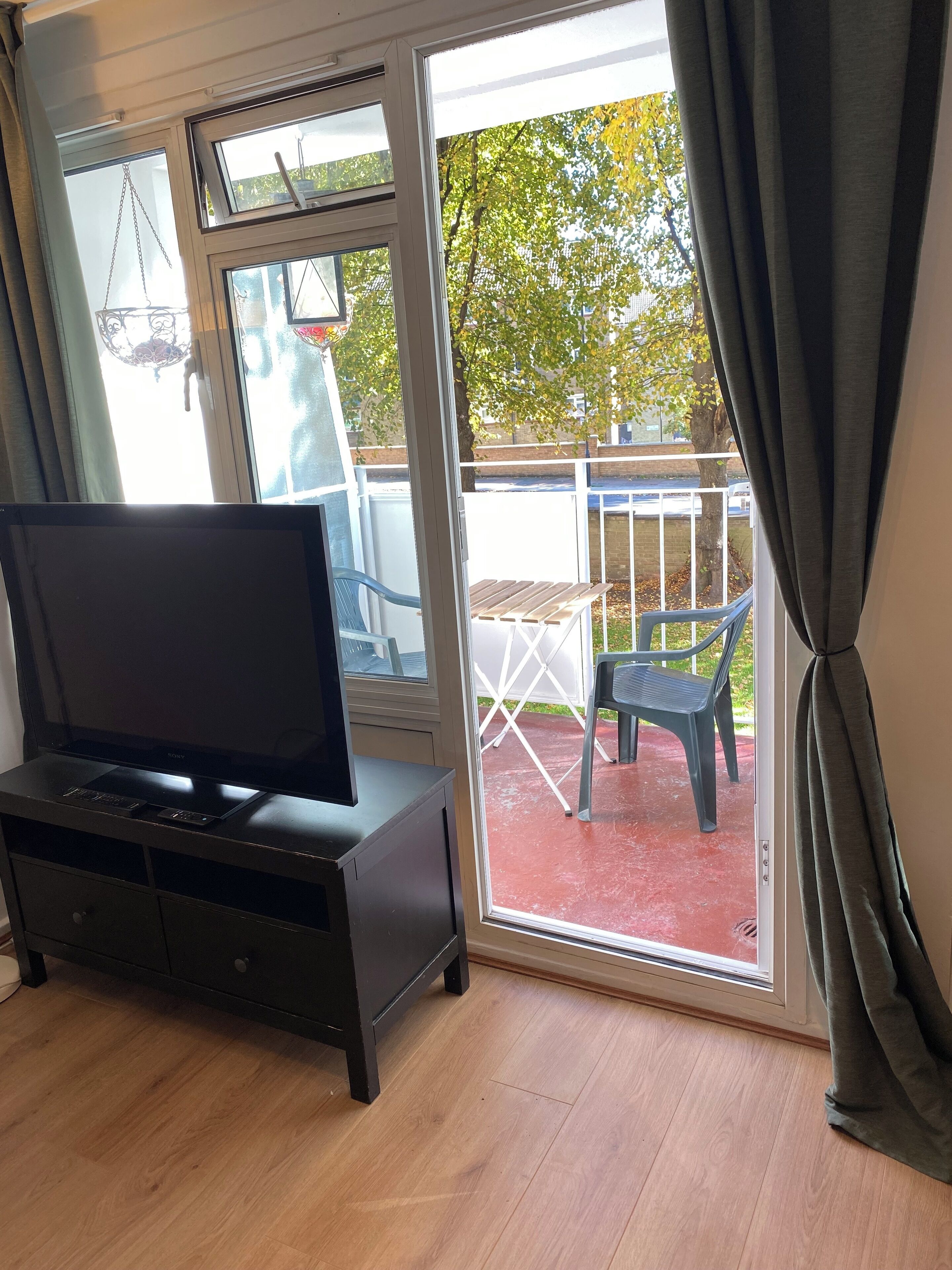 A room with a TV on a black stand, a sliding glass door leading to an outdoor area with a table and chairs, and a basketball hoop visible through the door.